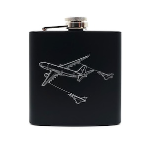 Könnte beinhalten: Schwarze Edelstahl-Taschenflasche mit silbernem Deckel und Klappverschluss. Die Flasche zeigt eine weiße Strichzeichnung eines Flugzeugs und zweier Kampfflugzeuge. Die Flasche ist rechteckig und hat eine glatte Oberfläche.
