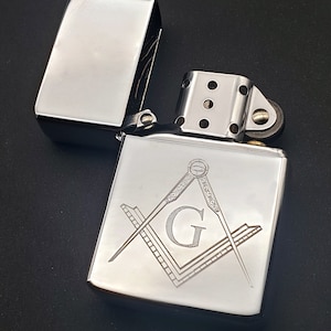 May include: A silver lighter with a Masonic symbol engraved on the front. The symbol features a compass and square with a 'G' in the centre.