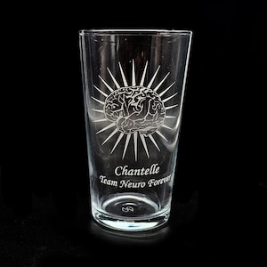 Puede incluir: Un vaso de pinta transparente con un diseño de cerebro blanco grabado, rodeado de líneas radiantes. El texto "Chantelle Team Neuro Forever" está grabado debajo. El vaso está sobre una superficie negra.