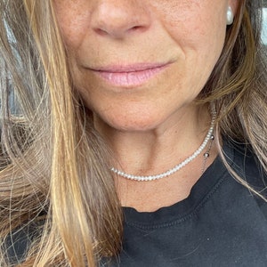 May include: A woman wearing a black top with a delicate pearl necklace. The necklace is a single strand of small, white pearls.