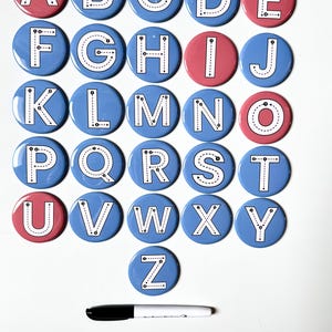 May include: A set of twenty-six circular buttons, each featuring a letter of the alphabet from A to Z. The letters are white with black dashed lines and directional arrows for tracing. The buttons are colored in blue or pink, accompanied by a black marker.