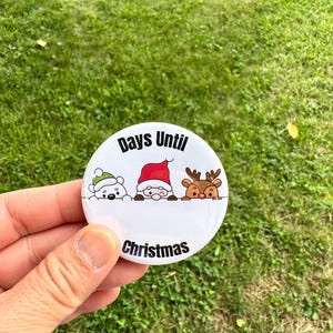 May include: A round white button featuring a cartoon illustration of a polar bear in a green hat, Santa Claus, and a reindeer peeking over a ledge. The text "Days Until" is printed above the characters and "Christmas" is printed below them.