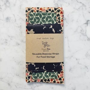 May include: A set of reusable beeswax wraps for food storage, featuring heart, geometric, and floral patterns. A brown label reads "Green Bee Wraps" and "Reusable Beeswax Wraps For Food Storage". The wraps are in small, medium, and large sizes.