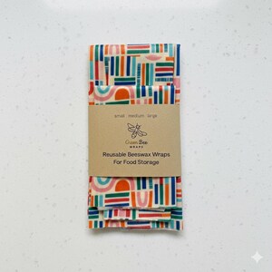 May include: A stack of reusable beeswax wraps for food storage. The wraps have a colorful geometric pattern in red, blue, green, and orange. A brown paper band with the text "Green Bee Wraps" and "Reusable Beeswax Wraps For Food Storage" is wrapped around the stack.