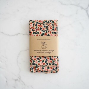May include: Folded reusable beeswax wraps for food storage. The wraps are beige with a pattern of red, green, and navy blue hearts. A brown paper band around the wraps reads "small medium large" and "Reusable Beeswax Wraps For Food Storage."