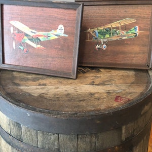 May include: Two framed vintage paintings of World War I era biplanes. The planes are painted in realistic detail with camouflage paint schemes. The frames are dark wood and the paintings are displayed on a wooden barrel.