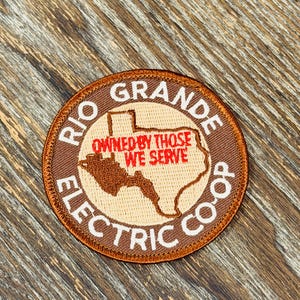 May include: A circular embroidered patch featuring a tan map of Texas centered on a brown background. The text RIO GRANDE ELECTRIC CO-OP is stitched in white around the border, while the red text OWNED BY THOSE WE SERVE is centered.