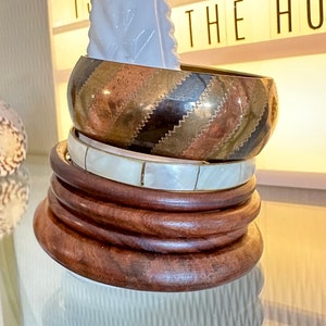 Peut inclure: Un ensemble de quatre bracelets empilés, comprenant un bracelet en bois, un bracelet en coquillage blanc et un bracelet en laiton à motif rayé. Les bracelets sont empilés les uns sur les autres et reposent sur une surface en verre.