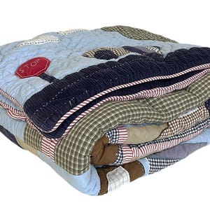 Puede incluir: Edredón patchwork plegado con un diseño de cielo azul, una sección de pana azul marino y una aplicación de señal de stop roja. El edredón presenta varios parches de tela a cuadros y lisos, y está ribeteado con un borde a rayas.