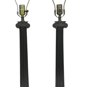 May include: Two bronze-colored table lamps with a classical column design. Each lamp features a fluted column base, a decorative capital, and a metal lamp shade holder. The lamps have a square base and a finial on top.