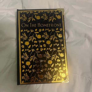 K&ouml;nnte beinhalten: Ein Hardcover-Buch mit dem Titel "On The Homefront" mit schwarzem Einband und goldenem Blumendesign. Das Buch hat goldene Kanten und Eckakzente. Das Cover zeigt ein sich wiederholendes Muster aus Bl&auml;ttern und Blumen.