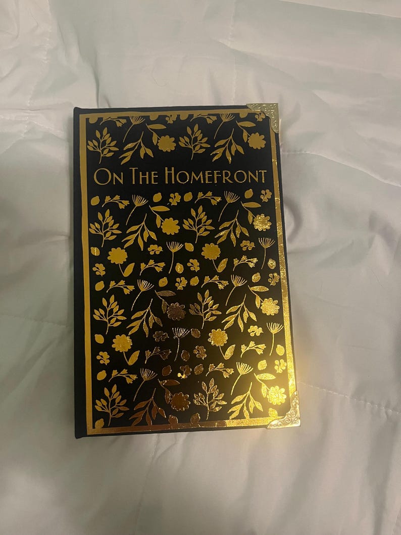K&ouml;nnte beinhalten: Ein Hardcover-Buch mit dem Titel "On The Homefront" mit schwarzem Einband und goldenem Blumendesign. Das Buch hat goldene Kanten und Eckakzente. Das Cover zeigt ein sich wiederholendes Muster aus Bl&auml;ttern und Blumen.