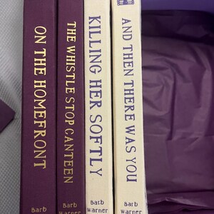 K&ouml;nnte beinhalten: Vier B&uuml;cher mit Titeln wie "On the Homefront", "The Whistle Stop Canteen", "Killing Her Softly" und "And Then There Was You". Die Buchr&uuml;cken sind lila und cremefarben mit goldfarbenen Buchstaben. Der Name des Autors ist unten auf jedem Buch aufgedruckt.
