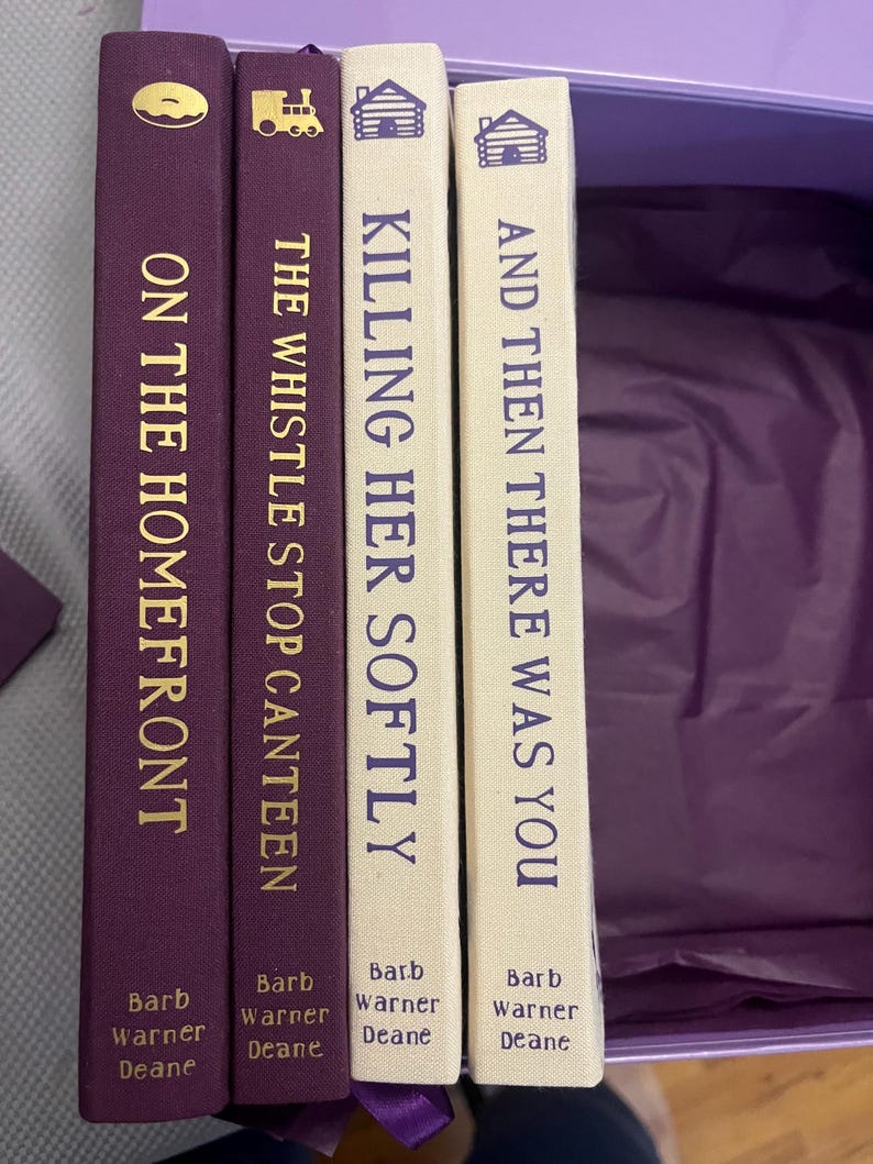 K&ouml;nnte beinhalten: Vier B&uuml;cher mit Titeln wie "On the Homefront", "The Whistle Stop Canteen", "Killing Her Softly" und "And Then There Was You". Die Buchr&uuml;cken sind lila und cremefarben mit goldfarbenen Buchstaben. Der Name des Autors ist unten auf jedem Buch aufgedruckt.
