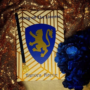 K&ouml;nnte beinhalten: Ein Buch mit dem Titel "Song of the Lioness" von Tamora Pierce. Das Cover zeigt ein blaues Schild mit einem goldenen L&ouml;wenemblem, vor einem cremefarbenen und goldenen geometrischen Muster. Ein blaues Lesezeichen und blaue Blumen sind ebenfalls zu sehen.