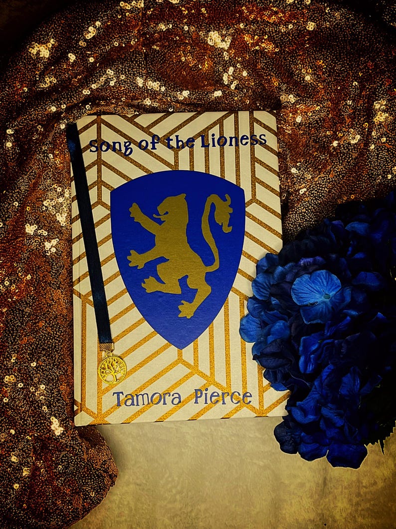K&ouml;nnte beinhalten: Ein Buch mit dem Titel "Song of the Lioness" von Tamora Pierce. Das Cover zeigt ein blaues Schild mit einem goldenen L&ouml;wenemblem, vor einem cremefarbenen und goldenen geometrischen Muster. Ein blaues Lesezeichen und blaue Blumen sind ebenfalls zu sehen.