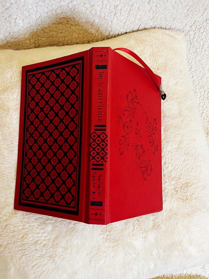 K&ouml;nnte beinhalten: Ein rotes Hardcover-Buch mit dem Titel "The Scarlet Letter" von Nathaniel Hawthorne. Das Buch hat ein dekoratives schwarz-rotes geometrisches Muster auf dem Einband und dem Buchr&uuml;cken. Ein rotes Lesezeichen mit Herzanh&auml;nger ist befestigt.