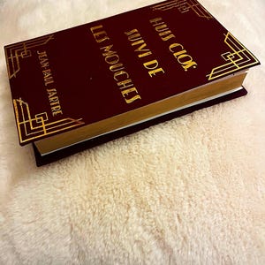 K&ouml;nnte beinhalten: Ein weinrotes Buch mit goldfarbenen Buchstaben und dekorativen Eckendesigns. Der Buchtitel lautet "Huis Clos Suivi de Les Mouches" von Jean-Paul Sartre. Das Buch hat goldfarbene Kanten und liegt auf einer flauschigen, cremefarbenen Oberfl&auml;che.