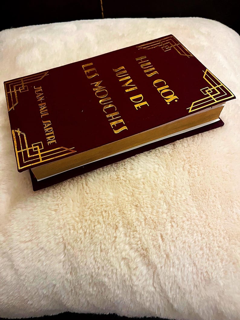 K&ouml;nnte beinhalten: Ein weinrotes Buch mit goldfarbenen Buchstaben und dekorativen Eckendesigns. Der Buchtitel lautet "Huis Clos Suivi de Les Mouches" von Jean-Paul Sartre. Das Buch hat goldfarbene Kanten und liegt auf einer flauschigen, cremefarbenen Oberfl&auml;che.