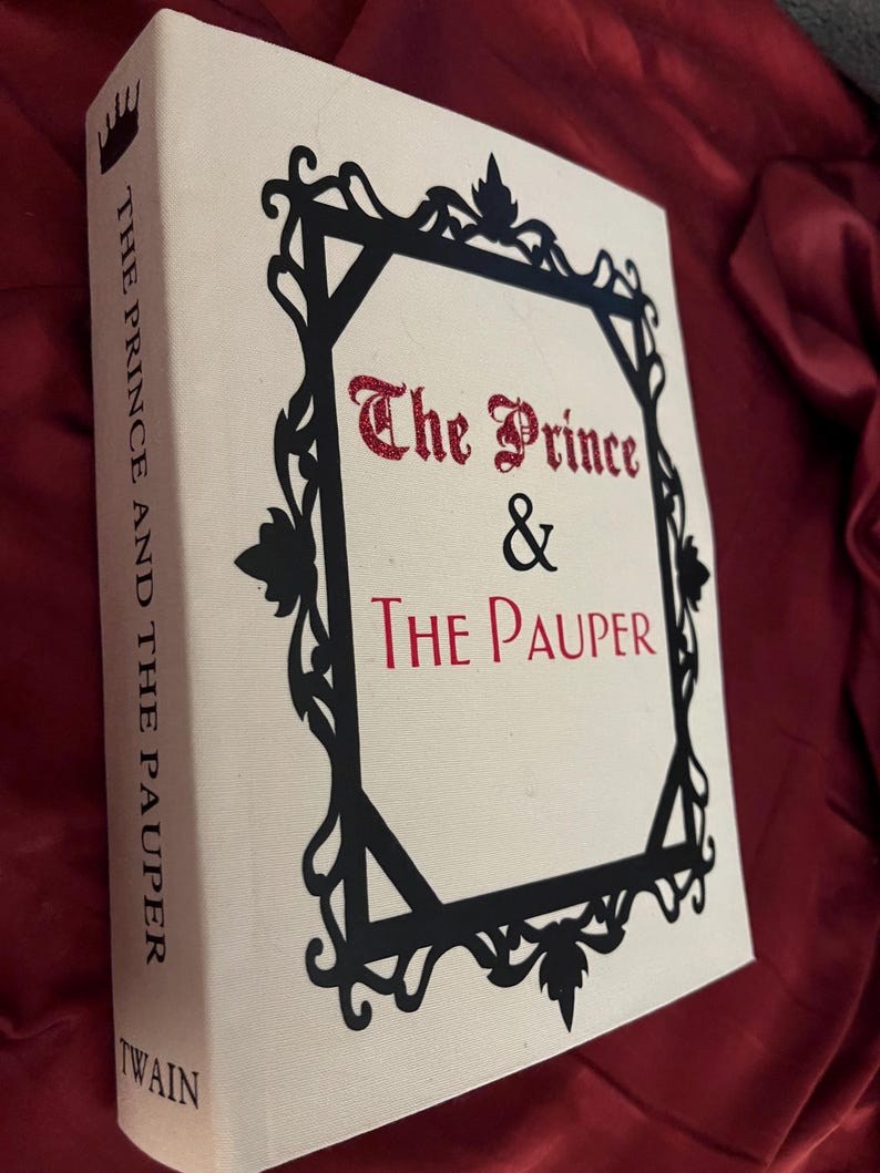 K&ouml;nnte beinhalten: Ein cremefarbenes Hardcover-Buch mit dem Titel "The Prince & The Pauper" von Twain. Das Buch hat einen schwarzen dekorativen Rahmen und roten Glitzerschriftzug. Der Buchr&uuml;cken zeigt den Titel und den Namen des Autors.