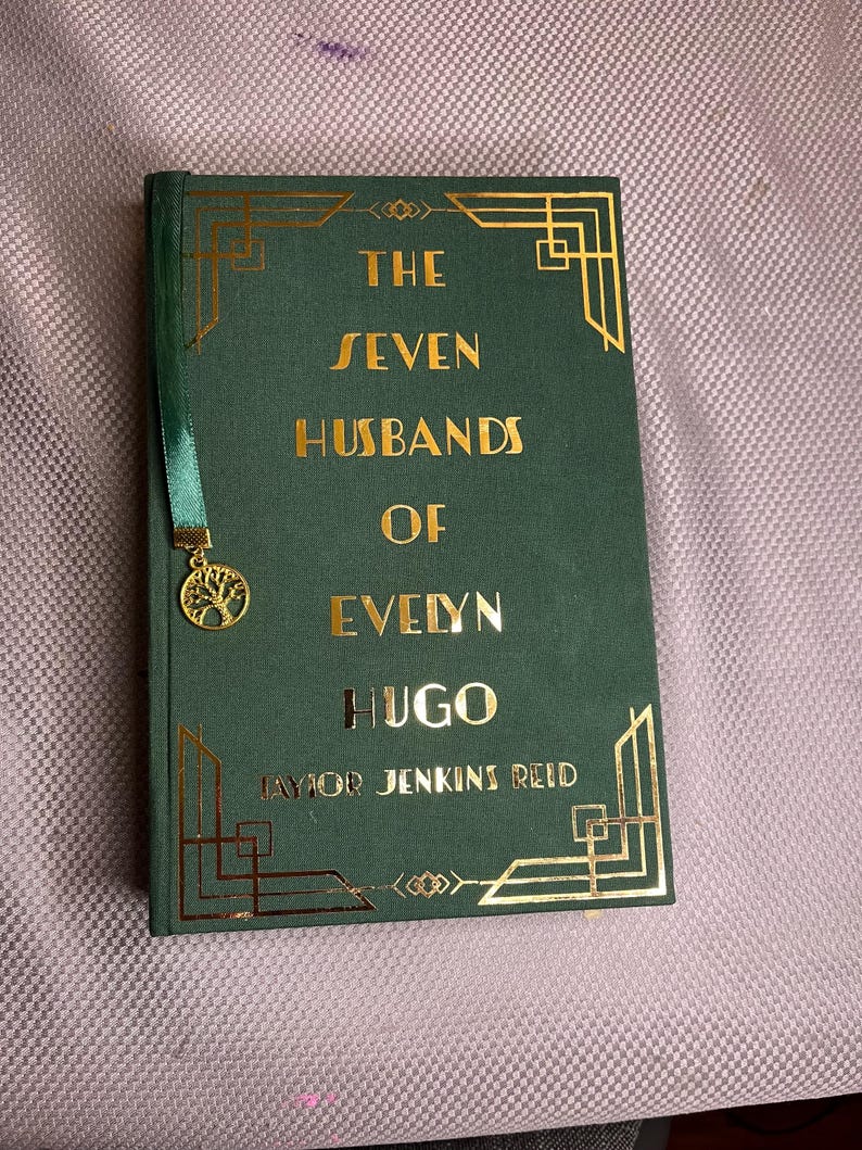 K&ouml;nnte beinhalten: Ein gr&uuml;nes Hardcover-Buch mit dem Titel "The Seven Husbands of Evelyn Hugo" mit goldfarbenen Buchstaben und Art-D&eacute;co-Akzenten. Ein gr&uuml;nes Lesezeichen mit goldenem Baum-Anh&auml;nger ist angebracht.