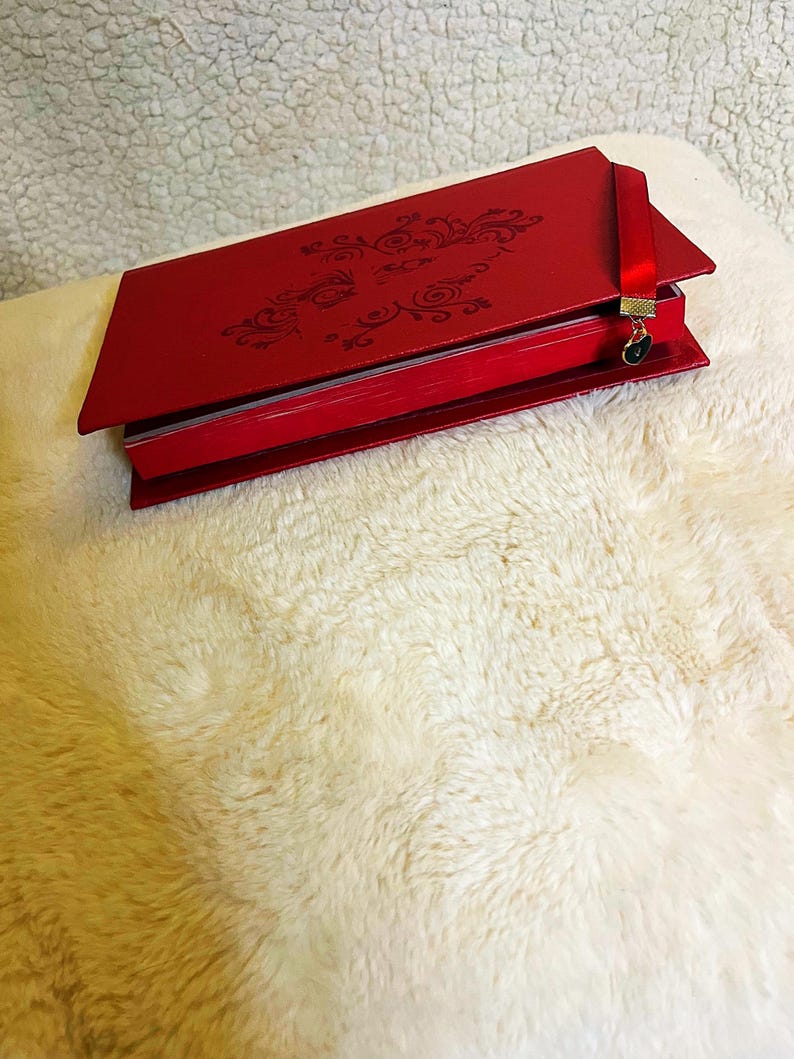 K&ouml;nnte beinhalten: Ein rotes Notizbuch mit einem verzierten Design auf dem Einband. Das Notizbuch hat ein rotes Lesezeichenband mit einem kleinen Anh&auml;nger. Das Notizbuch ist ge&ouml;ffnet und zeigt die Seiten im Inneren. Es liegt auf einer flauschigen, wei&szlig;en Oberfl&auml;che.
