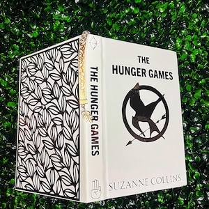 May include: A white book titled "The Hunger Games" with a black mockingjay symbol and the author's name, Suzanne Collins. The book has a black and white leaf pattern on the side and a gold bookmark.
