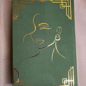 K&ouml;nnte beinhalten: Ein gr&uuml;nes Notizbuch mit goldenem Art-D&eacute;co-Design. Der Einband zeigt eine goldene Umrisszeichnung eines Frauengesichts und geometrische Eckakzente. Oben ist ein goldenes Schleifenlesezeichen zu sehen. Das Notizbuch ist rechteckig und geschlossen.