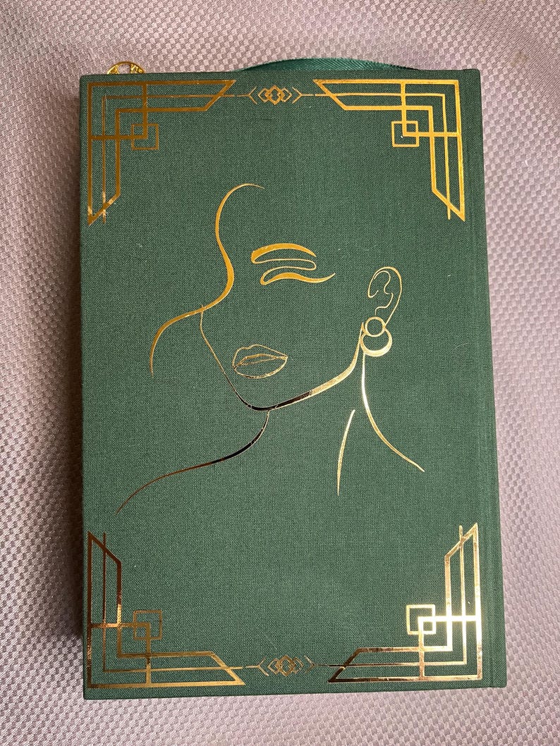 K&ouml;nnte beinhalten: Ein gr&uuml;nes Notizbuch mit goldenem Art-D&eacute;co-Design. Der Einband zeigt eine goldene Umrisszeichnung eines Frauengesichts und geometrische Eckakzente. Oben ist ein goldenes Schleifenlesezeichen zu sehen. Das Notizbuch ist rechteckig und geschlossen.