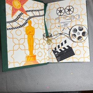K&ouml;nnte beinhalten: Offenes Buch mit gr&uuml;nem Einband. Die Seiten zeigen Illustrationen zum Thema Film, darunter eine goldene Oscar-Statue, ein roter Stern, Filmstreifen, eine Filmkamera und eine Filmklappe. Der Hintergrund hat ein geometrisches Muster.