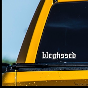 May include: A yellow vehicle with a black window and a white decal that reads "bleghsssed" in a gothic font. The image is a close-up of the vehicle's rear window and body.