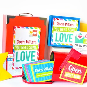 May include: A red metal mailbox with a flap that says "Open Right Now!" and a smaller red metal box with a flap that says "Open When You Are Feeling Happy". There is a larger red metal box with a flap that says "Open When You Need Some Love".