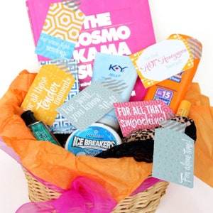 May include: A gift basket filled with various items including a pink book titled The Cosmo Kama Sutra, a bottle of K-Y Jelly, sunscreen, Ice Breakers mints, and a leopard print garment, all accented with colorful tags featuring text like For all those tender moments.