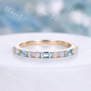 May include: A gold band ring with alternating light blue and iridescent gemstones. The ring features a series of rectangular and round-cut stones, creating a delicate and elegant design. The ring is set against a soft, blurred background.