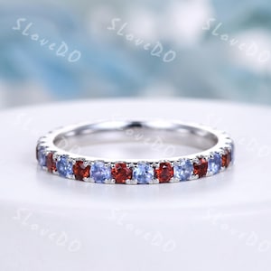 Puede incluir: Un anillo de banda plateado con piedras preciosas rojas y azules alternas. El anillo está engastado con pequeñas piedras preciosas redondas que rodean toda la banda. El anillo se muestra sobre una superficie blanca.