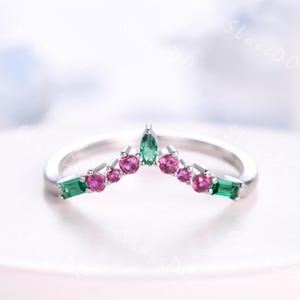 Könnte beinhalten: Ein silberner Ring mit Kronen-Design. Der Ring hat einen zentralen grünen Edelstein, flankiert von kleineren rosa Edelsteinen und grünen rechteckigen Edelsteinen. Die Edelsteine sind auf einem polierten Silberband gefasst.