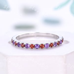 May include: A silver ring with a band of alternating purple and red gemstones. The gemstones are evenly spaced around the ring, creating a continuous pattern. The ring is set against a soft, blurred background.