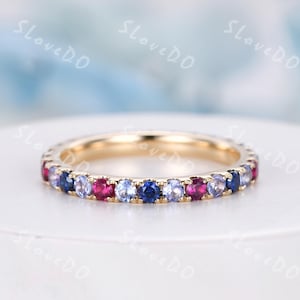 May include: A gold band ring featuring a continuous row of alternating red, blue, and purple gemstones. The ring is set on a white surface with a blurred background.