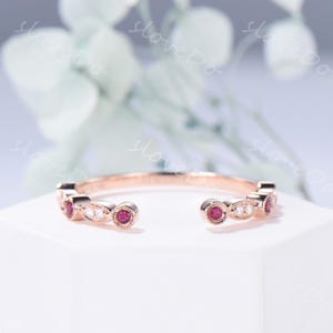 May include: Rose gold ring with a delicate design. The ring features alternating round red gemstones and small white gemstones. The ring is set on a white surface with a blurred background.