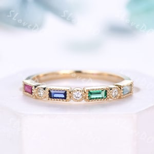 Puede incluir: Un anillo de banda de oro con una fila de piedras preciosas de colores. El anillo presenta una piedra preciosa rosa, azul, verde y azul claro, cada una engastada en un engaste de bisel de oro. El anillo está acentuado con pequeños diamantes redondos.