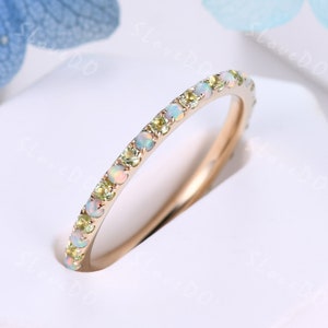 May include: A delicate gold band ring with a row of small, round, white opals and green gemstones. The ring is set on a white background.