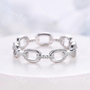 May include: A silver ring with a chain link design. The ring features oval and rectangular links, some of which are encrusted with small, clear gemstones. The ring is set against a soft, white background.