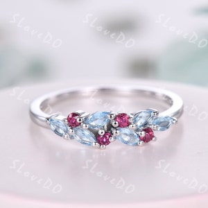 May include: A silver ring with a delicate design featuring blue and red gemstones. The ring has a cluster of marquise-shaped blue stones and round red stones.