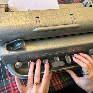 May include: A silver Perkins Brailler typewriter with a sheet of paper with braille text. The typewriter has a textured surface and is on a plaid surface. The image shows a person's hands using the typewriter.