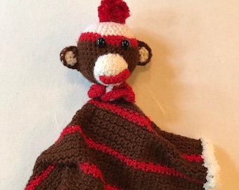 Puede incluir: Una manta de seguridad de mono de calcetín hecha a mano. El mono tiene una cabeza marrón con detalles blancos y rojos, ojos de botón negros y un pompón rojo en su gorro. La manta es marrón con rayas rojas y un ribete blanco.