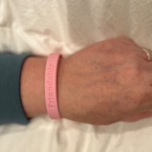 May include: A light pink silicone bracelet with the word "Friendship" embossed on it. The bracelet is worn on a wrist. The background is a blue shirt and white fabric.