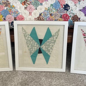 May include: Three square white frames containing fabric art pieces. Each piece features a central black square with two triangular fabric wings extending outward. The wings display floral patterns in yellow, teal, and pink on a cream background.