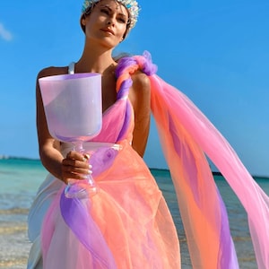 May include: A woman wearing a jeweled crown and holding a large, lavender-colored crystal singing bowl. She is draped in flowing fabric in shades of pink, orange, and purple. The background is a clear blue sky and ocean.