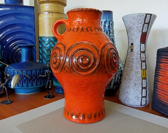 Puede incluir: Jarrón de cerámica naranja con un diseño de espiral marrón y un asa. El jarrón está sobre una superficie gris claro.