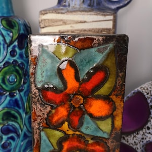 May include: A rectangular ceramic vase with a floral design. The vase features a vibrant orange flower with teal and yellow leaves. The background includes other ceramic vases with blue and white patterns.